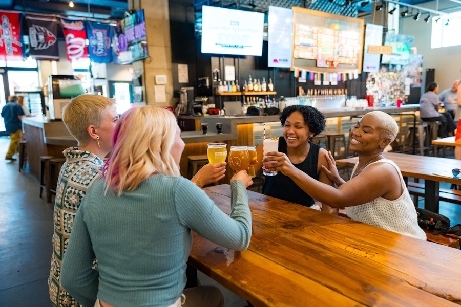 Two couples smile and clink their beers together at a brewery. 
