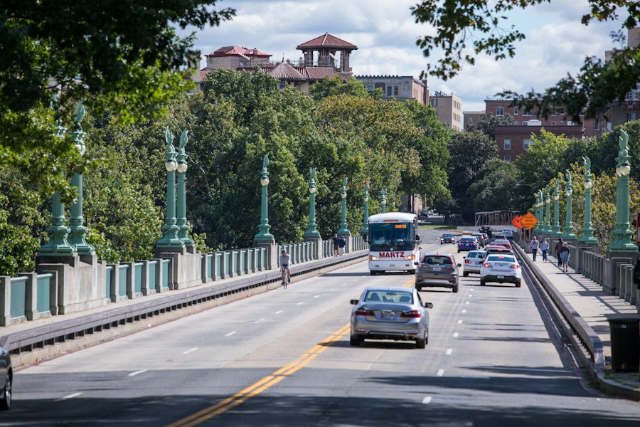 Vehicles and cyclists traverse a tree-lined city bridge under a partly cloudy sky, with historic buildings in the background.