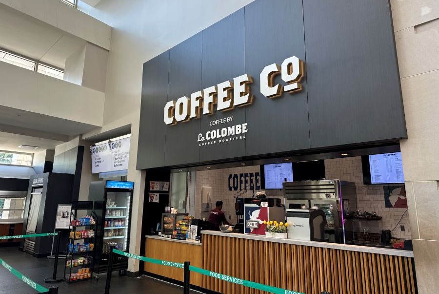 A modern coffee kiosk named 'Coffee Co. by La Colombe' inside a building, with a counter, digital menus, and customers being served.