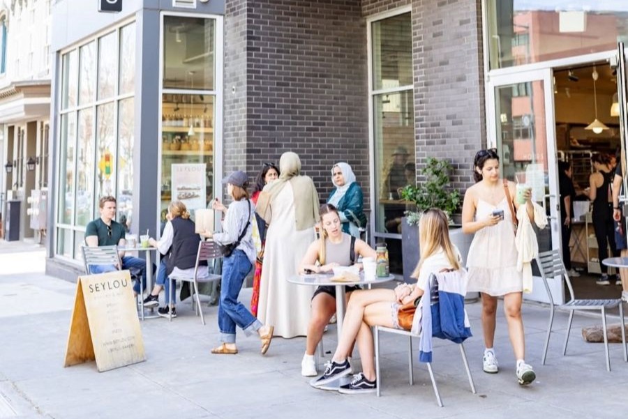 People gather and relax outside Seylou Bakery, enjoying food and drinks at sidewalk tables on a sunny day.