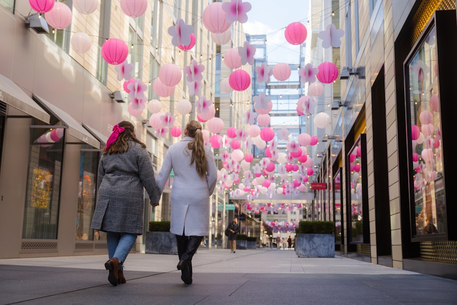 A couple in winter coats walks through modern storefronts with pink lanterns hanging above. 