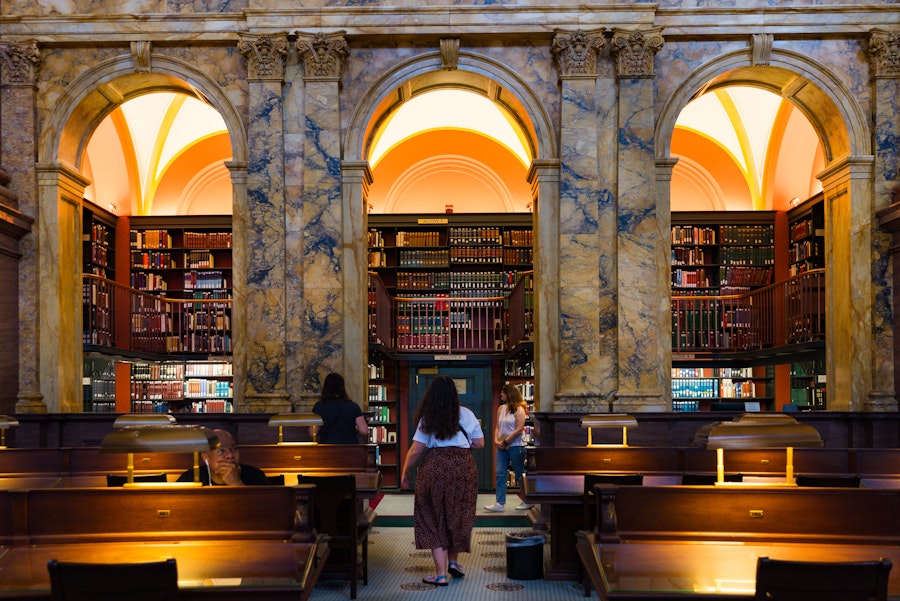 Patrons read and study inside a richly decorated reading room with arched bookshelves and glowing lamps.