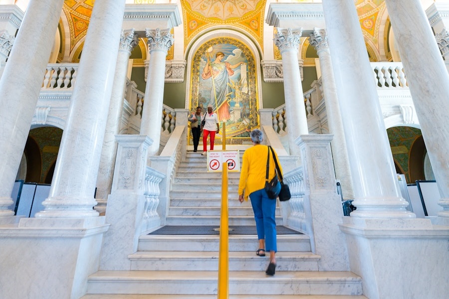 A visitor ascends the grand marble staircase toward a vibrant mosaic mural inside the Library of Congress.
