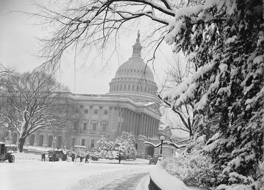 Black-and-white photo of the U.S. Capitol surrounded by snow-covered trees with workers clearing the street.
