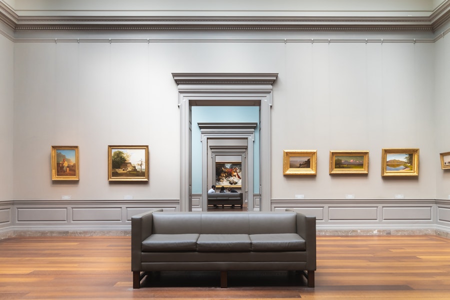 View down a series of connected gallery rooms with paintings on the walls and gray couches in the center.