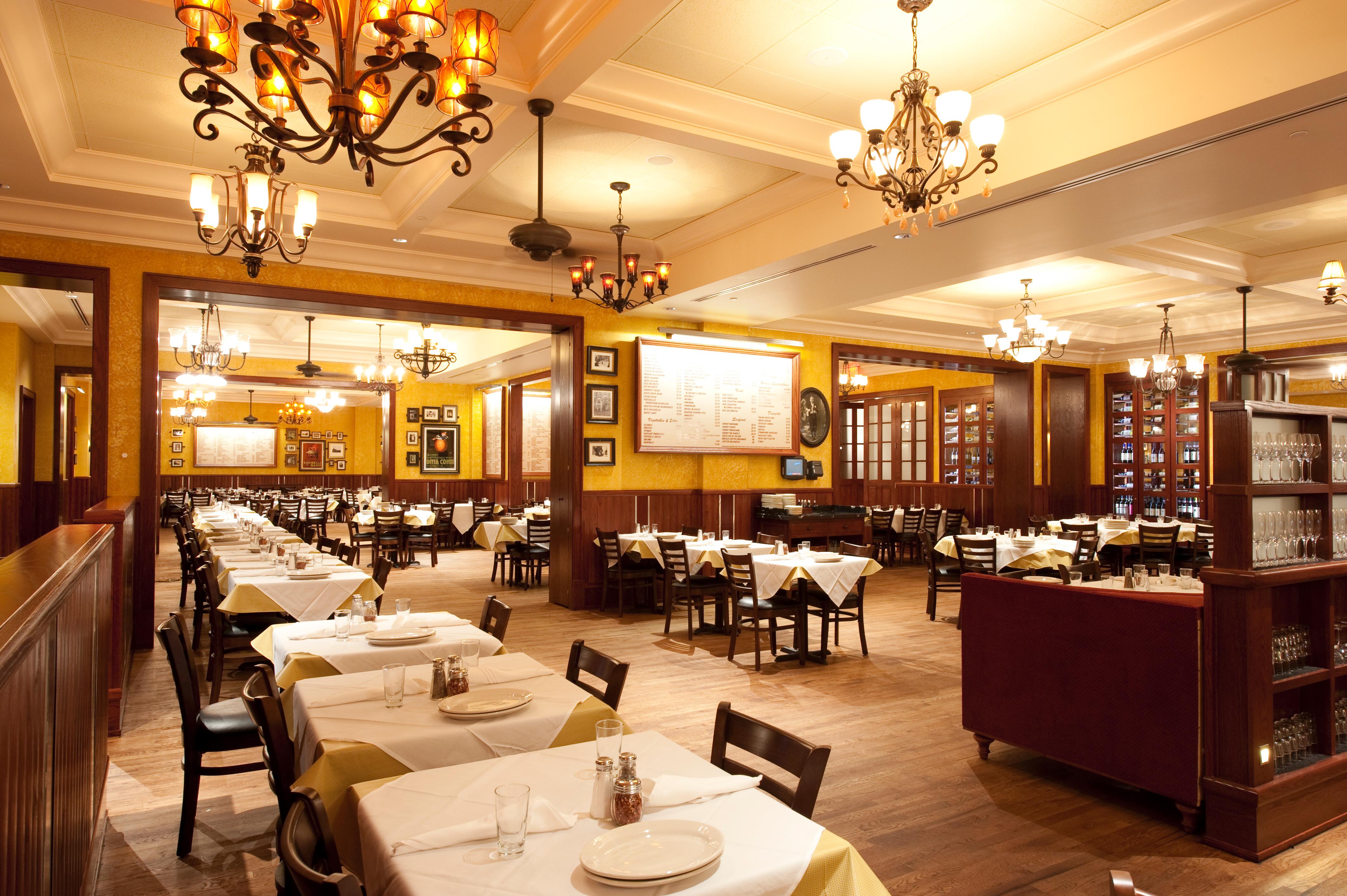 A warmly lit, spacious restaurant dining room features neatly set tables with white tablecloths, wooden chairs, chandeliers overhead, and walls lined with framed art and wine displays.