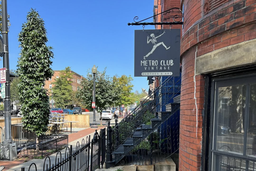 The sign for Metro Club Vintage, a shop selling clothes and art, hangs outside a brick building on a sunny street.