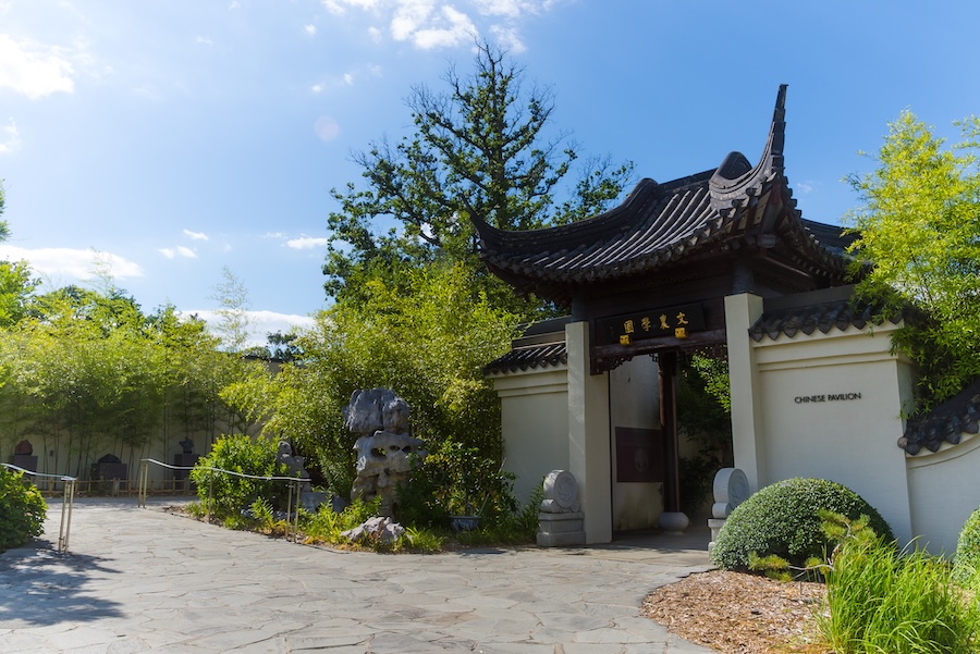 The Chinese Pavilion at the U.S. National Arboretum features a traditional curved roof surrounded by lush greenery and rock elements.