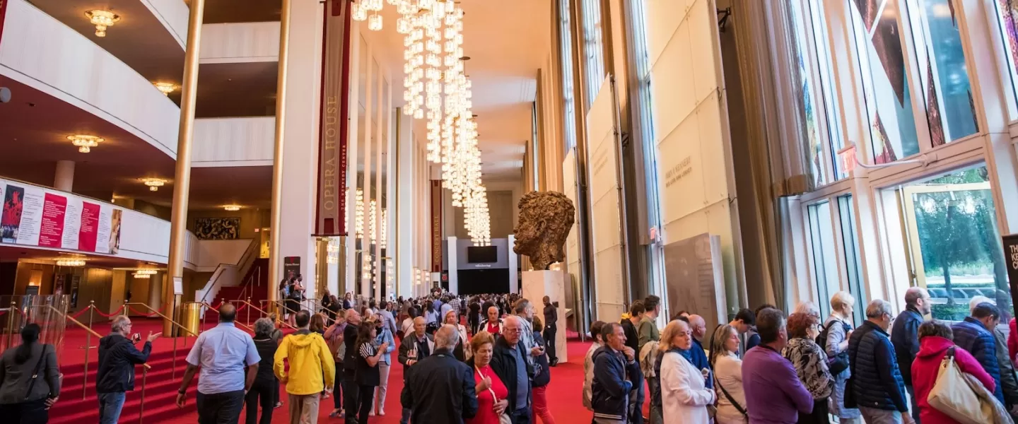 A crowded lobby inside the Kennedy Center, with people walking past a large sculpture and chandeliers under a high ceiling.