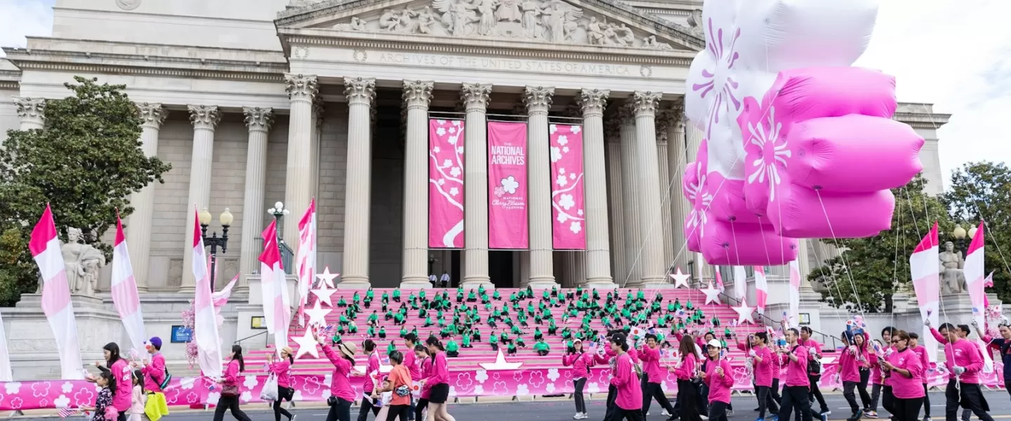 Participants in bright pink attire march with banners and balloons during the National Cherry Blossom Festival parade in front of the National Archives in Washington, DC.