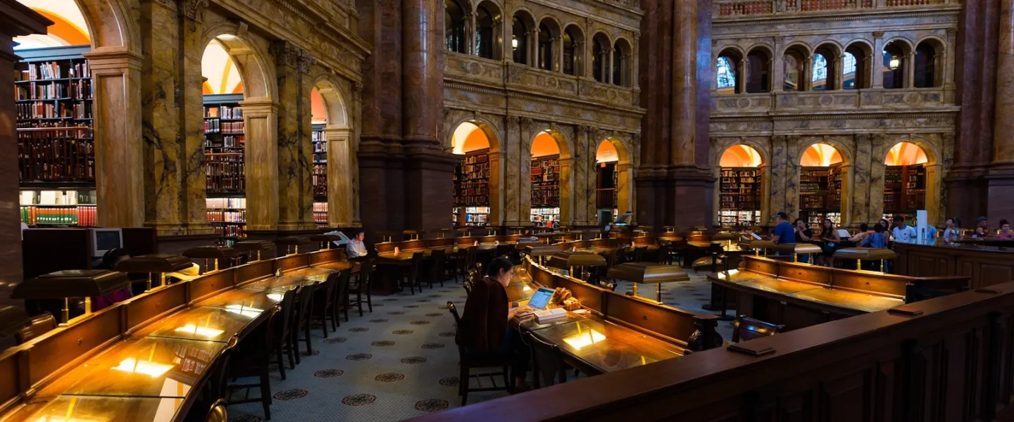 Visitors study and explore the grand marble reading room at the Library of Congress surrounded by arched alcoves and bookshelves.