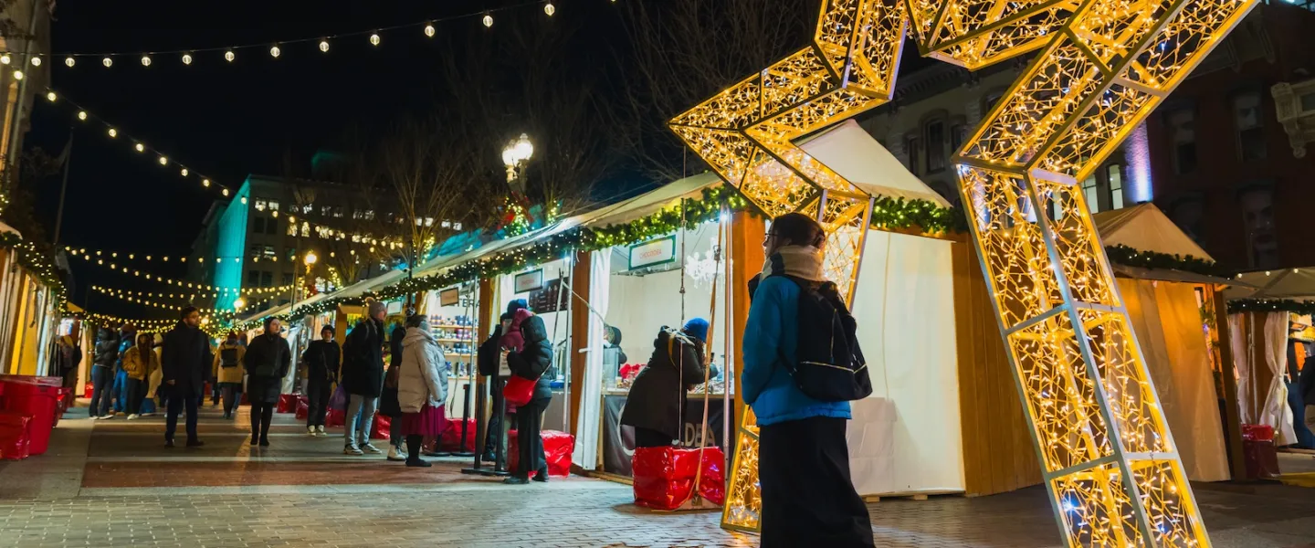Shoppers browse vendor stalls lined with string lights and greenery at the Downtown Holiday Market in Washington, DC, with a large illuminated star installation marking the entrance.