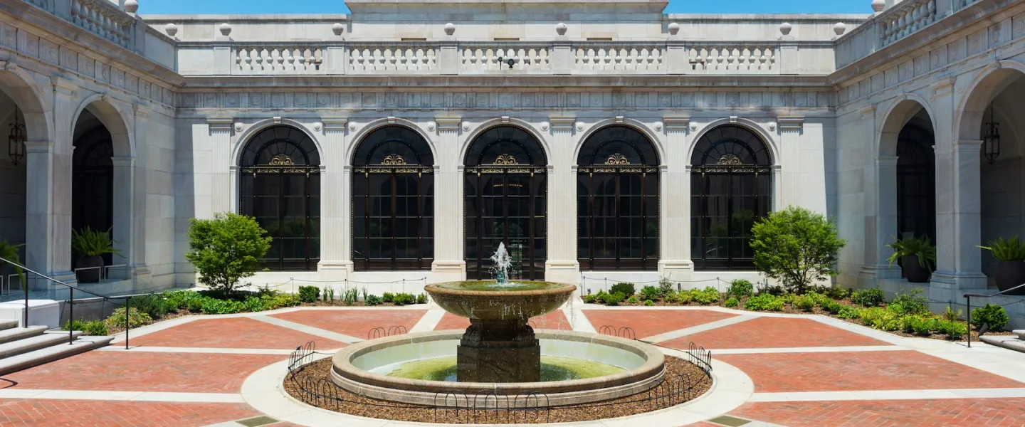 A round fountain sits at the center of a bright brick courtyard surrounded by white stone arches and large windows in Washington, DC.