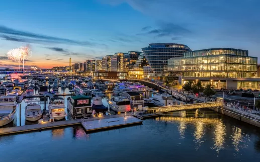  A vibrant waterfront scene at dusk with illuminated modern buildings, boats docked at the marina, and fireworks lighting up the sky in the distance.