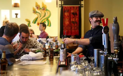 Bartender with a mustache serves drinks to patrons at a busy bar, decorated with vibrant artwork in the background.