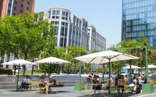 A lively outdoor plaza with people sitting under white umbrellas surrounded by modern glass buildings and leafy green trees on a sunny day.