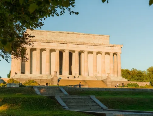 The Lincoln Memorial at sunset framed by trees with golden light on the monument’s columns.