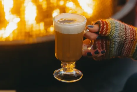 A hand in a sweater holds out a glass mug filled with boozy hot chocolate in front of an outdoor fireplace.