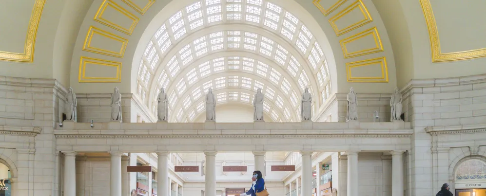  A woman in a blue coat and brown boots walks with a blue suitcase through the grand, arched interior of a spacious train station adorned with statues and large windows.