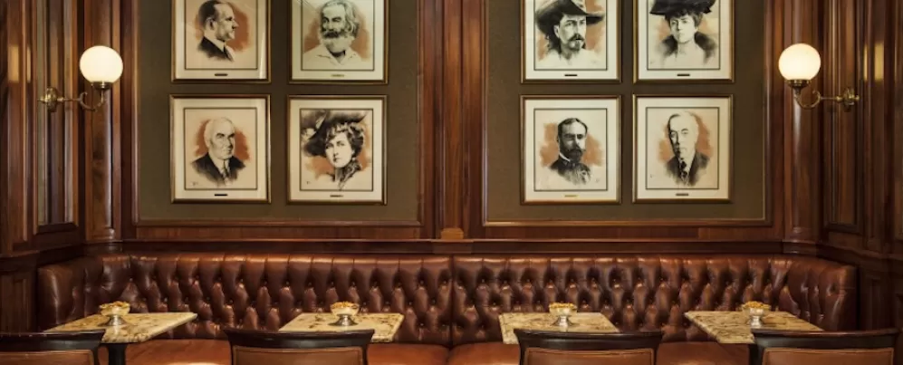 A cozy seating area featuring tufted leather banquettes, small marble tables, and portraits of notable historical figures adorning the walls in a warm wooden setting.