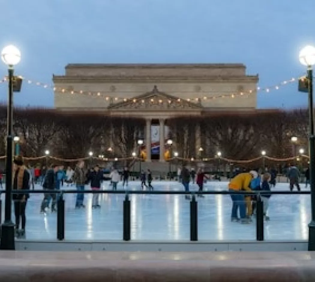 People ice skating at the National Gallery of Art Sculpture Garden rink during the evening, with string lights overhead and the museum building in the background.